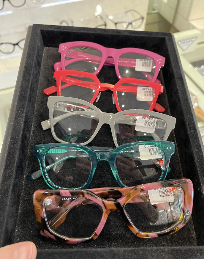 a black tray holds five eyeglass pairs in pink, red, gray, teal, and tortoiseshell and each has a price tag