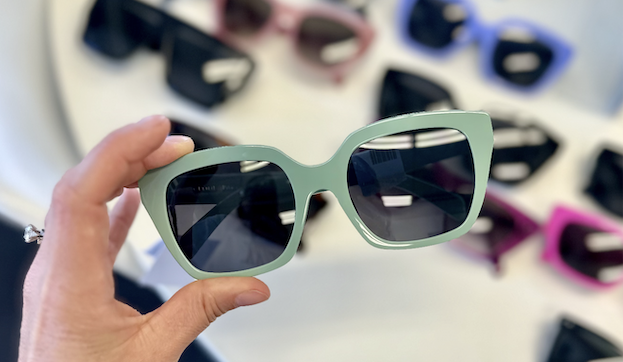 a hand holds oversized square sunglasses with green frames and dark lenses, displayed among other sunglasses